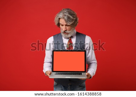 Amazed elderly gray-haired mustache bearded man in shirt vest tie posing isolated on red background. People lifestyle concept. Mock up copy space. Holding laptop pc computer with blank empty screen