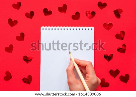 White blank notepad with woman hand holding pen on red background with red shiny hearts confetti