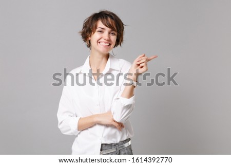 Smiling pretty young business woman in white shirt posing isolated on grey wall background studio portrait. Achievement career wealth business concept. Mock up copy space. Pointing index finger aside