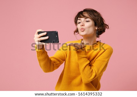 Pretty young brunette woman girl in yellow sweater posing isolated on pastel pink background. People lifestyle concept. Mock up copy space. Doing selfie shot on mobile phone, blowing sending air kiss