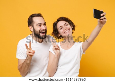 Smiling couple friends bearded guy girl in white t-shirts isolated on yellow orange background. People lifestyle concept. Mock up copy space. Doing selfie shot on mobile phone, showing victory sign