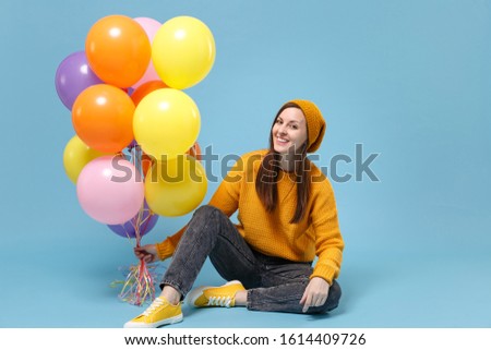 Smiling young woman girl in sweater and hat posing isolated on blue background. Birthday holiday party people emotions concept. Mock up copy space. Celebrating holding colorful air balloons sitting