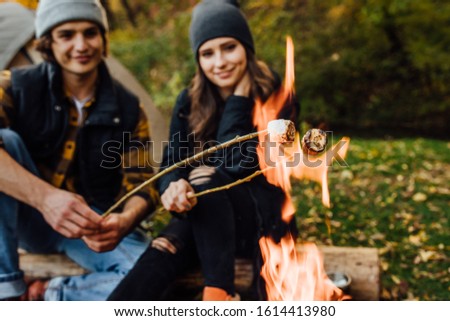 Close up photo of roasting marshmallows over the fire near tent in camping. Focus on fire. Young loving couple of tourists relaxing near the fire in the nature.