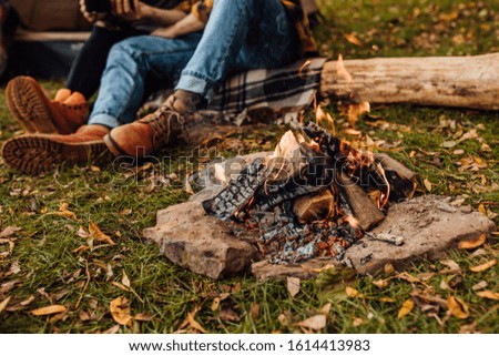 Close up photo of bonfire in the forest near tent. Happy couple on the log near fire.