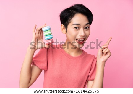 Young Asian girl over isolated pink background holding colorful French macarons and pointing up a great idea