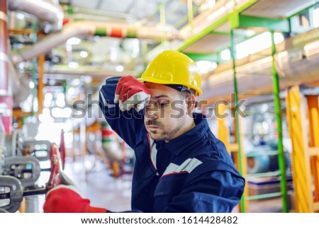 Hardworking handsome caucasian plant worker in protective suit and with helmet on head trying to screw valve while wiping sweat. Royalty-Free Stock Photo #1614428482