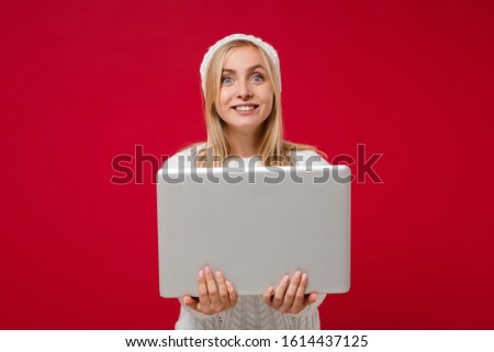 Smiling young woman in white sweater hat isolated on red wall background in studio. Healthy fashion lifestyle people sincere emotions, cold season concept. Mock up copy space. Hold laptop pc computer