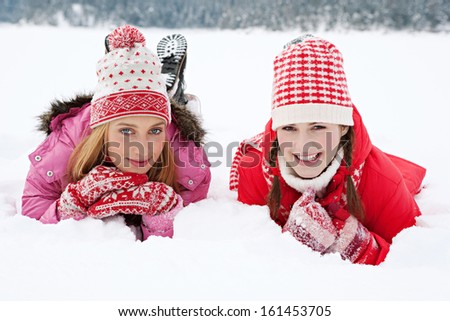 Two joyful and attractive women friends laying down together on white snow while on a vacation trip to the skiing mountains, being playful during a cold winter day, outdoors.