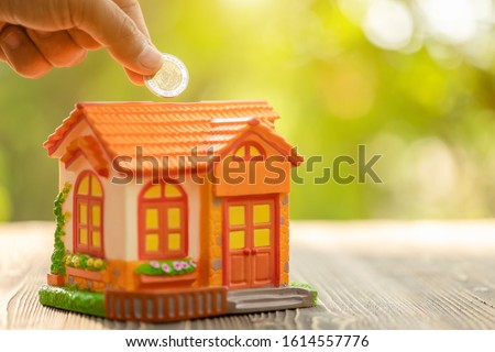 Close up hand put the coin to piggy bank (Home Model) on green nature blur background. Money savings concept