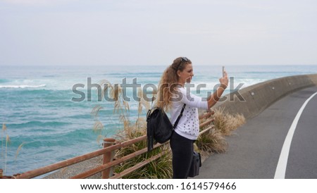 young beautiful curly-haired girl on the road makes a photo on a mobile phone. road by the ocean in Japan Aburatsu