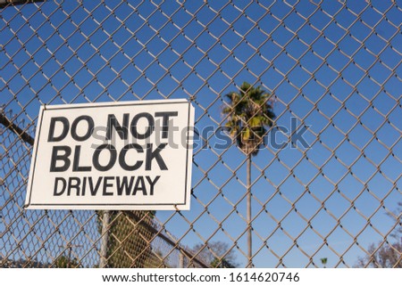 "Do not block driveway" sign on a chain link fence with a tall palm tree and blue sky in the background in Los Angeles, USA