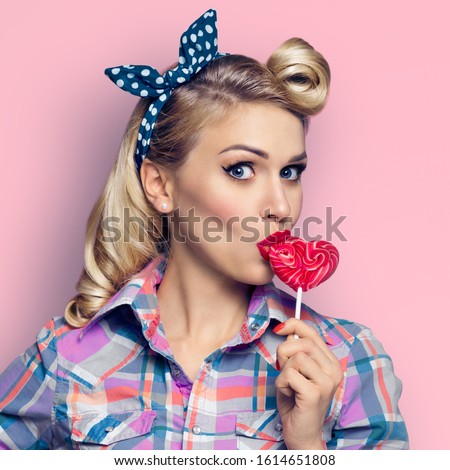 Surprised woman eating heart shape candy lollipop. Girl in pin up. Blond model - retro fashion and vintage. Pink background. Square composition. 