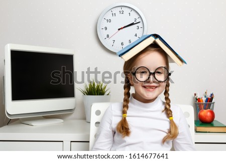 A cheerful little girl student with glasses sits in a room next to a table with a book on her head. Education and school concept.