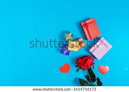 Six gift boxes with red, pink, dark blue, orange, yellow and green colors placed on a blue background, located in the center of the picture. There are red rose and heart at the bottom of the picture