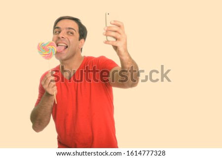Happy handsome Persian man taking selfie while licking heart shaped lollipop