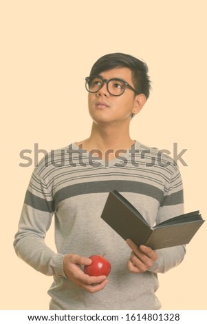 Studio shot of young handsome Asian man holding book and red apple while thinking