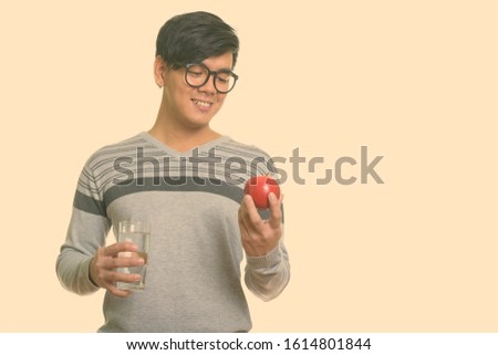 Young happy Asian man smiling and looking at red apple while holding glass of water
