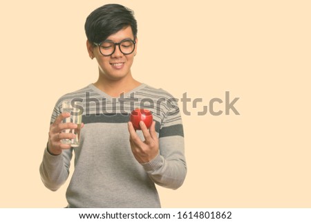 Young happy Asian man smiling and looking at red apple while holding glass of water