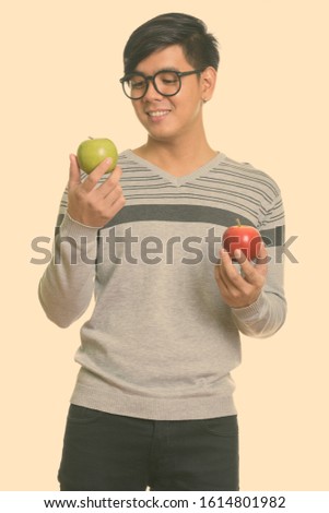 Studio shot of young happy Asian man smiling while choosing between red and green apple