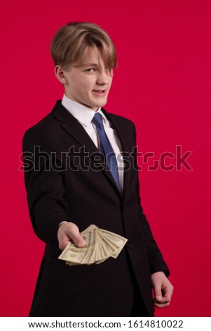 Teen boy in a black jacket holds a bundle of dollars banknotes in his hands and poses on a red background