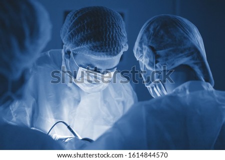Three surgeons during abdominal surgery. Blue uniforms, latex gloves, medical instruments. Spot lighting.
