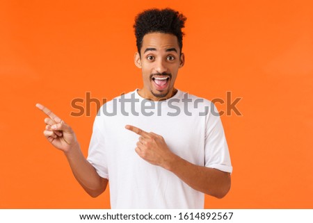 You see, check it out. Amused and excited cheerful african-american hipster guy in white t-shirt, pointing upper left corner and smiling, suggest awesome place, introduce product, orange background
