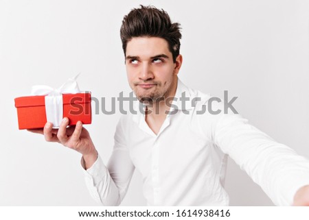 Funny caucasian man in shirt holding present. Studio shot of emotional brunette guy celebrating valentine's day.