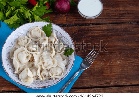 Boiled dumplings with feathers of green onions. In the background are greens, red peppers and bay leaves. In a transparent bowl sour cream.