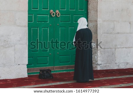 Muslim religious woman in long dress and covered head standing against the massive green door of mosque and praying. Back view of woman standing near the entrance of the Dome of the Rock in Jerusalem