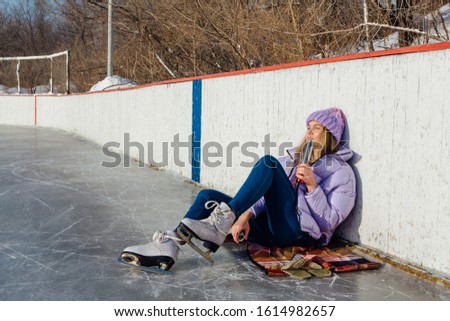 Lovely young woman relaxing after riding ice skates and drinking hot drink from termo pot on the ice rink. Girl sitting on ice and smiling.
