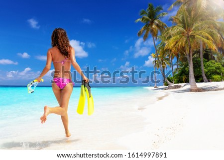 A attractive woman in bikini and with snorkeling gear in her hands runs on a tropical paradise beach with turquoise  sea, fine sand and coconut palm trees, Maldives islands