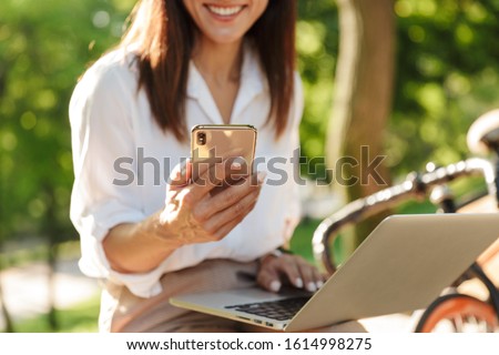 Cropped image of an attractive young woman relaxing on the bench at the city park, working on laptop computer, using mobile phone