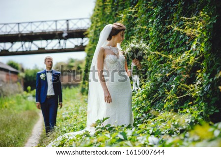 Beautiful bride with blond hair in stylish wedding dress with lace holds wedding bouquet of white flowers and greens, the groom on background. Wedding photo session outdoors