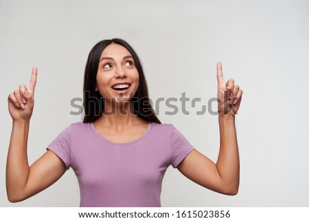 Studio shot of excited young dark haired female with casual hairstyle pointing upwards with raised forefingers and smiling positively, standing against white background