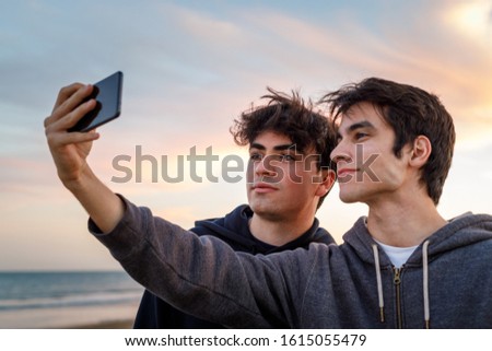 Two young friends on the beach at sunset having their photo taken with their mobile phone
