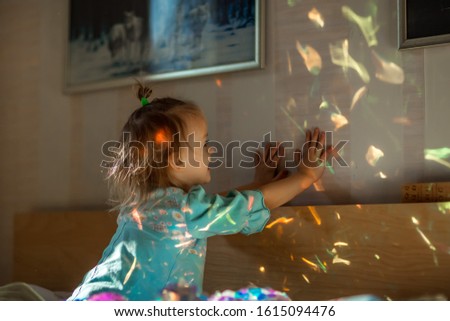 Little cute laughing girl in blue pajamas playing with sunlight spots made with a sparkling dress in bedroom on a sunny morning.