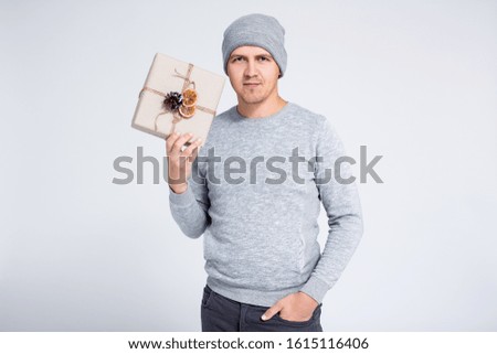 portrait of cheerful young man in warm winter clothes posing with gift box over gray background
