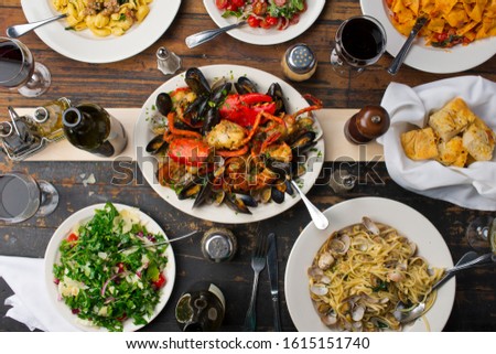 Italian dinner table with assortment of dishes. Mussels, pastas, pizzas, baked clams, octopus, whole fish, garlic bread and olive oil, meats, cheeses, olives, lobster and wine. 