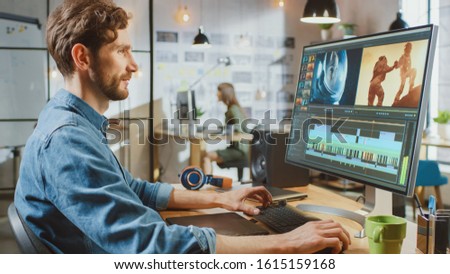 Male Video Editor with Beard and Jeans Shirt Works with Footage on His Personal Computer with Big Display. He Works in a Cool Office Loft. Other Female Creative Colleague Works in the Background.