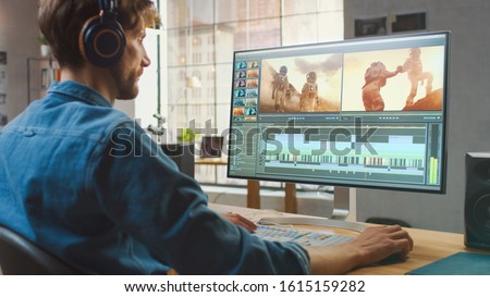 Male Video Editor in Headphones Working with Footages on His Personal Computer with Big Display. He Works in a Cool Office Loft. Creative Man Wears a Jeans Shirt. Royalty-Free Stock Photo #1615159282