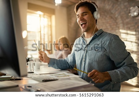 Happy freelance worker having fun while working at office desk and listening music over headphones. His colleagues are in the background.  Royalty-Free Stock Photo #1615291063