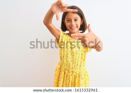 Young beautiful child girl wearing yellow floral dress standing over isolated white background smiling making frame with hands and fingers with happy face. Creativity and photography concept.