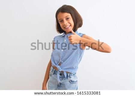 Young beautiful child girl wearing blue casual shirt standing over isolated white background doing happy thumbs up gesture with hand. Approving expression looking at the camera with showing success.