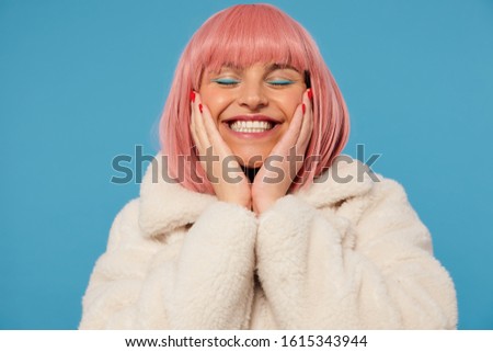 Pleased good looking young cheerful pink haired woman holding her face with raised palms while standing over blue background, smiling happily with closed eyes, dressed in fancy clothes