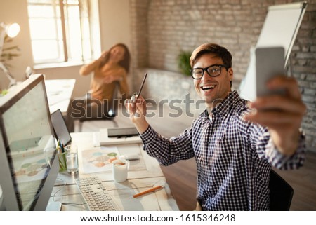 Young happy freelance worker using smart phone and taking selfie with his female coworker in the office. 