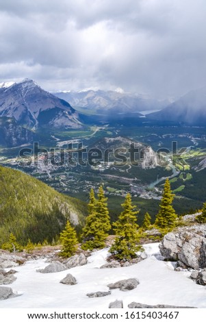 Snow covered mountain side with scattered pine trees in front of the view towards the snow capped rocky mountains, forest and town of Banff on a cloudy day.