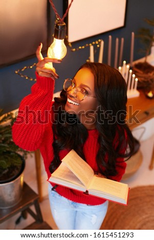 Girl in a red sweater and a book in hand poses in the Christmas interior at home. Happy Girl African American holding a New Year garland with his hand.