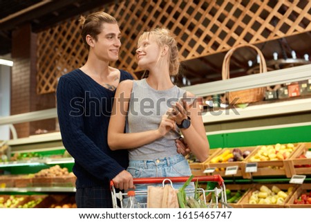 Young couple at the supermarket doing daily shopping standing with cart hugging holding notebook with shopping list looking at each other smiling happy Royalty-Free Stock Photo #1615465747