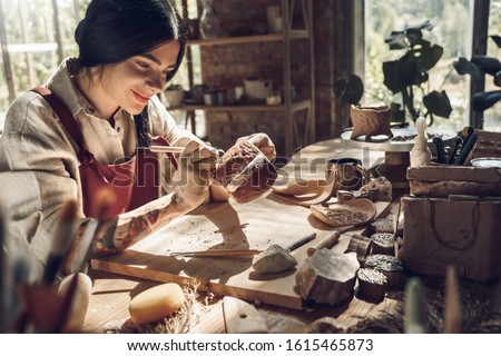Young woman wearing apron making pottery indoors at creative studio craftsperson concept sitting at table using modeling tool creating pattern on cup in the dark smiling cheerful close-up Royalty-Free Stock Photo #1615465873