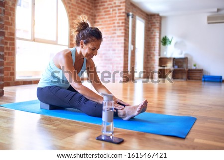 Middle age beautiful sportwoman smiling happy. Sitting on mat practicing yoga doing seated forward fold pose at gym